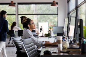 Confident young African American businesswoman smiles while using a desktop computer. She is working in an open office concept.